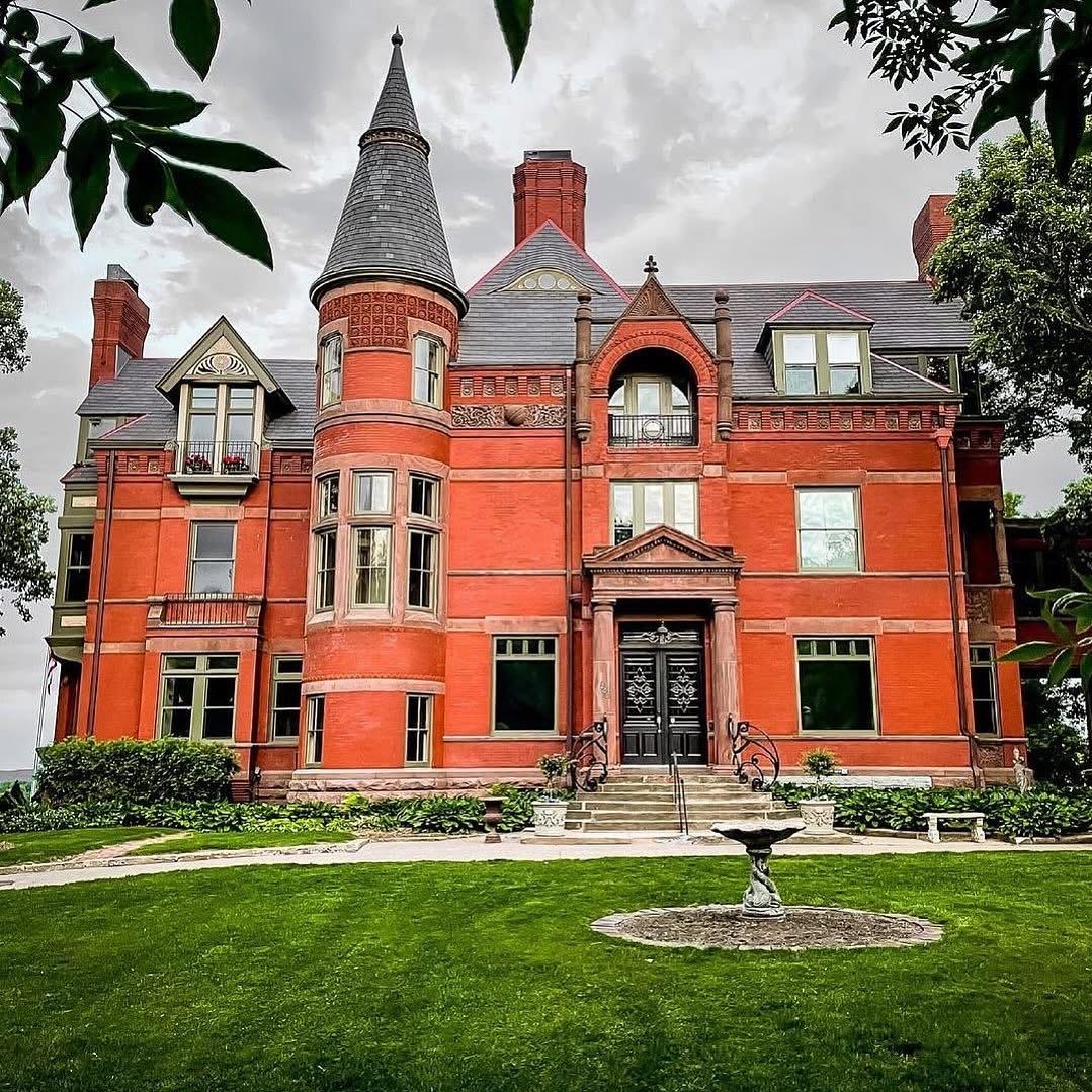 Red brick Victorian mansion with turret and ornate details on green lawn under cloudy sky