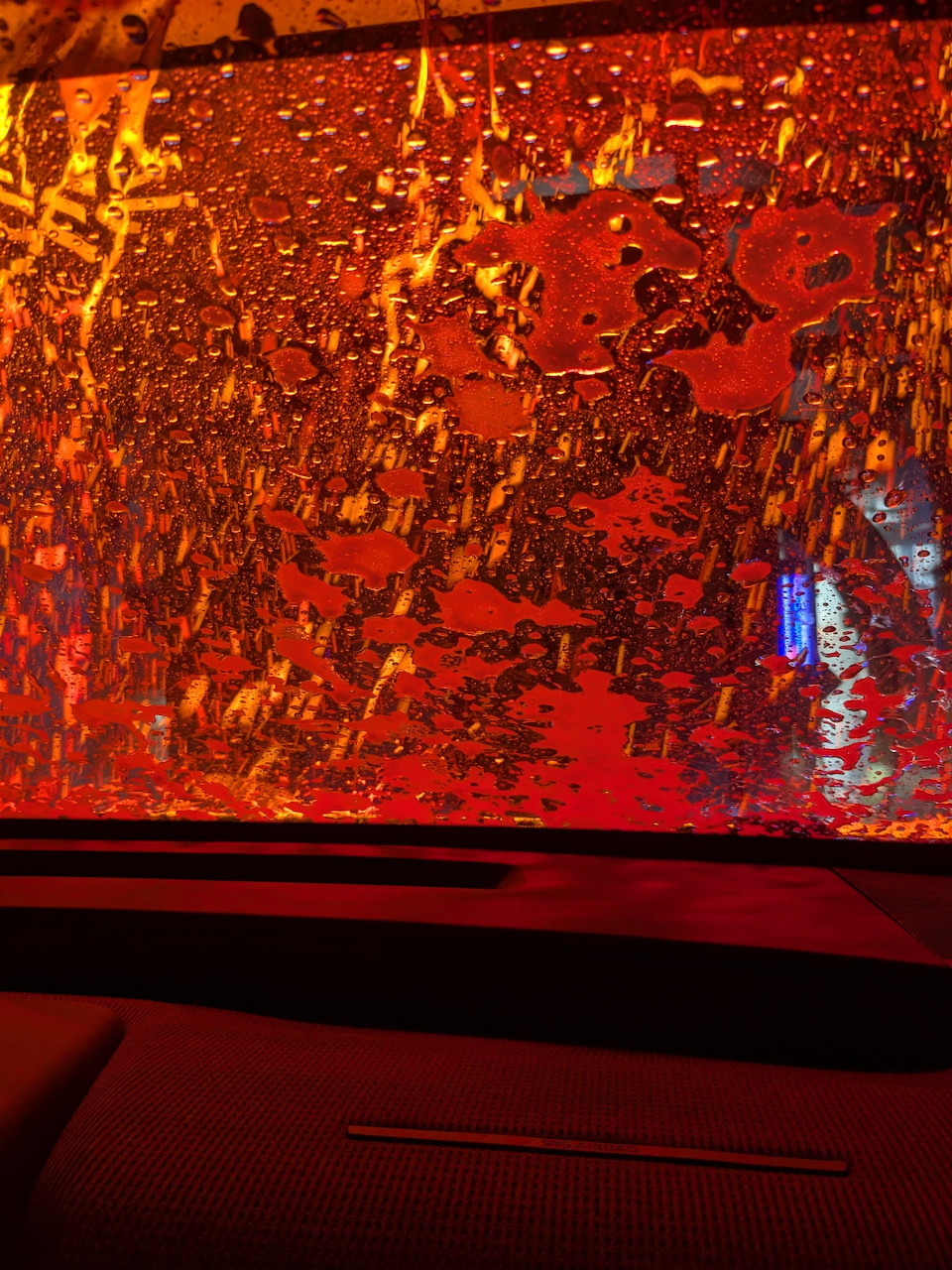 Wet car windshield with red and orange lights in a car wash, seen above a dark dashboard