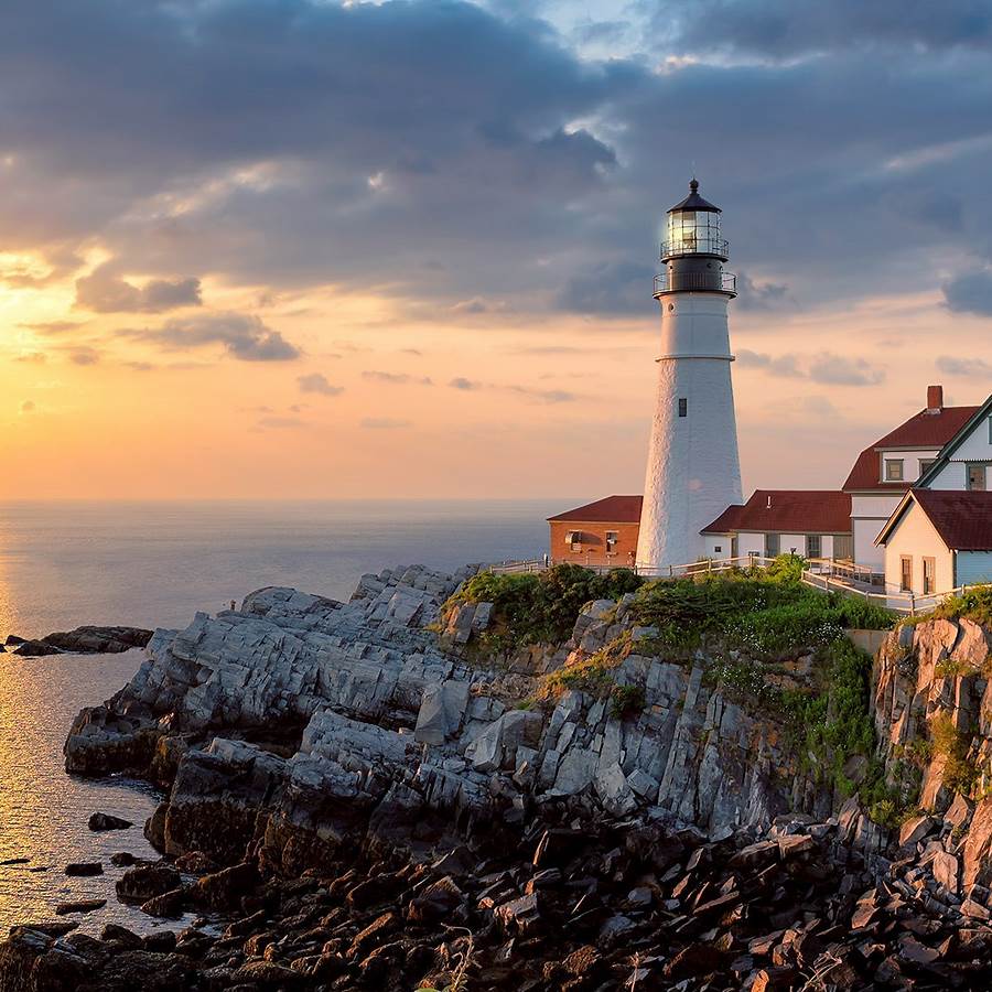 White lighthouse on rocky Maine coast at sunset with dramatic cloudy sky