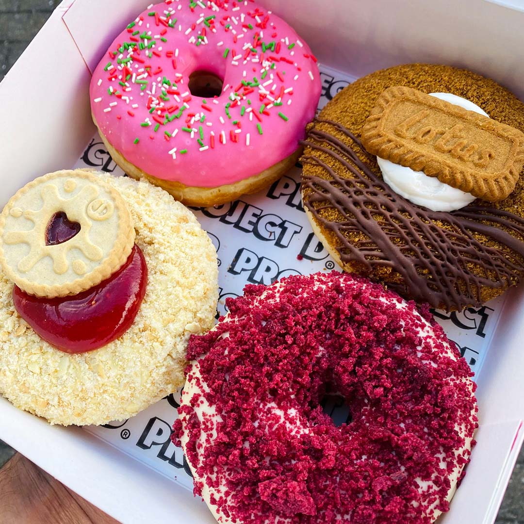 Box containing pink frosted donut, red velvet donut, sandwich cookies, and heart-shaped cookie with jam center