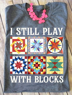 Gray t-shirt featuring colorful quilt block designs and text I Still Play With Blocks with pink necklace