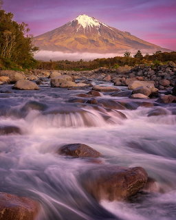 Snow-capped mountain beyond a rocky river with flowing water at dusk