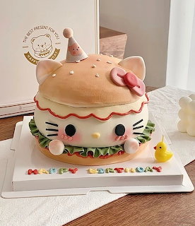 Hello Kitty burger-shaped cake on a white tray with colorful candy letters and a small yellow chick topper