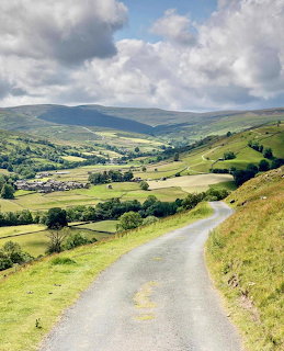 Winding road leading down into a green valley with hills and a village in the Yorkshire Dales