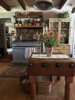 Rustic kitchen with wooden island, blue vintage cabinets, open shelves, exposed ceiling beams, and floral arrangement