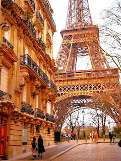 Eifel Tower seen from a Paris street with ornate Haussmann building on the left and pedestrians