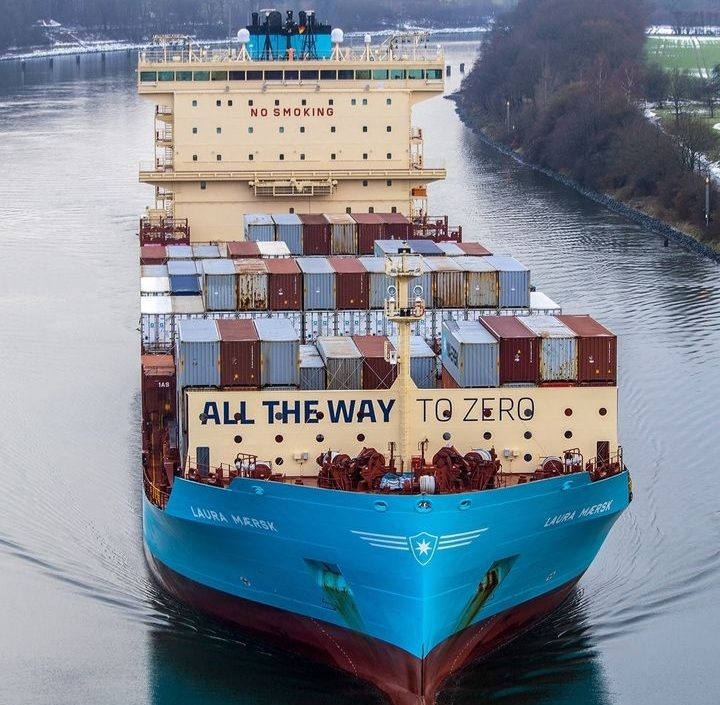 Blue container ship viewed from the front, carrying stacked cargo containers on a canal