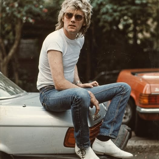 Roger Taylor sitting on a silver car wearing jeans, white T-shirt, and sunglasses