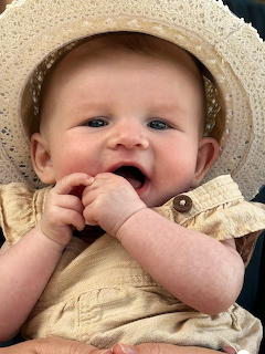 Smiling baby wearing a light sunhat and beige outfit with hands near the mouth