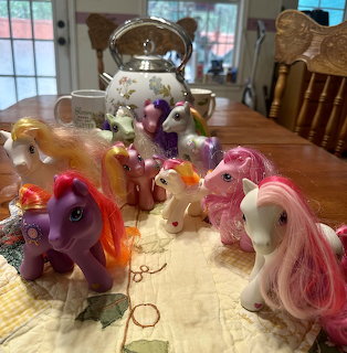 My Little Pony toys in pink, purple, and white gathered around a glass teapot on a kitchen table