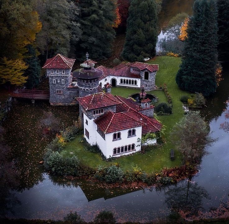 Small castle-like house with red roofs on a grassy island surrounded by water and trees