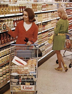 Two women in 1960s fashion shopping in a vintage supermarket with stocked shelves and a cart