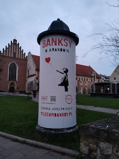 Cylindrical Banksy exhibition sign on a lawn in Krakow with historic buildings behind it