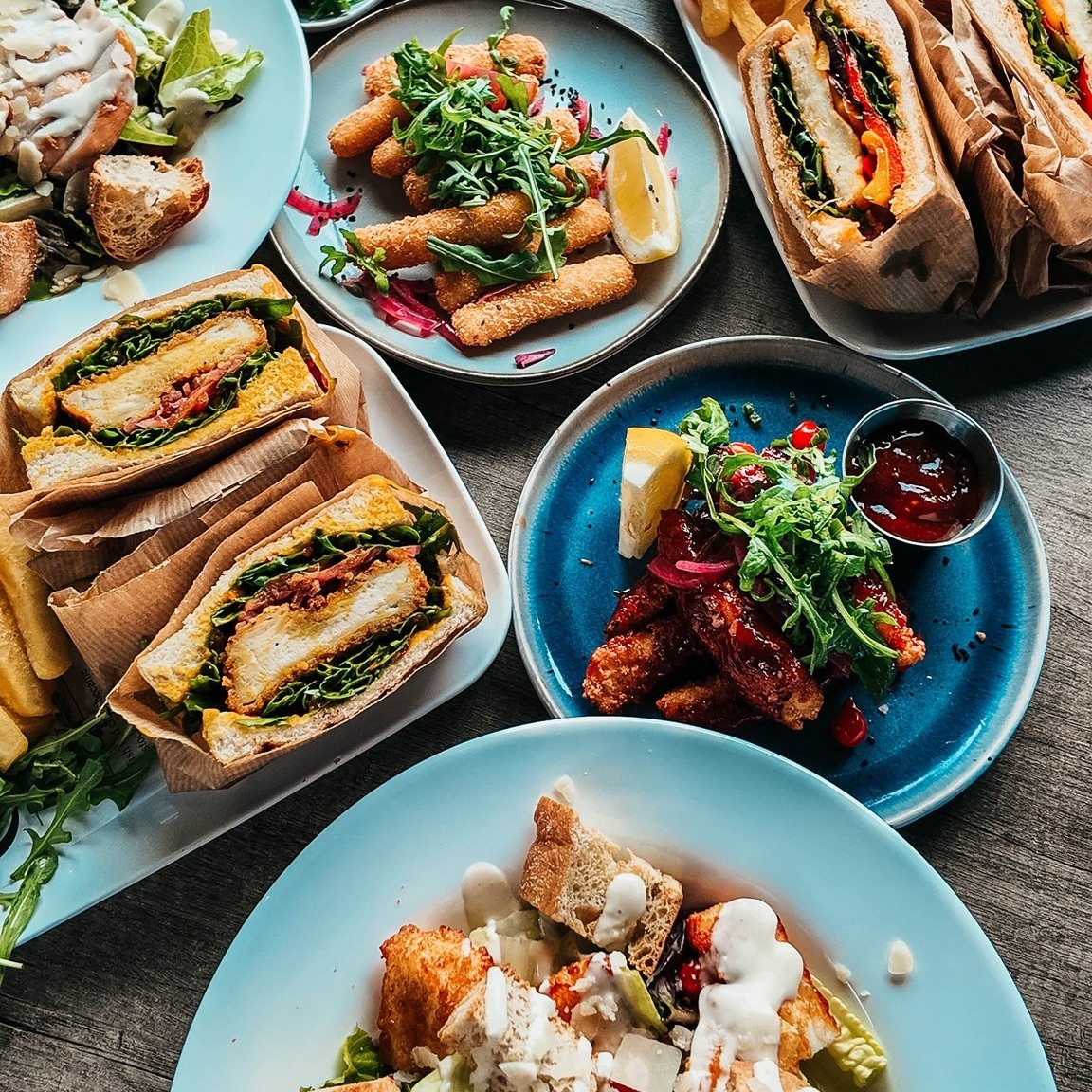 Overhead shot of various gourmet dishes on colorful plates including spring rolls, grilled meat, and fresh vegetables