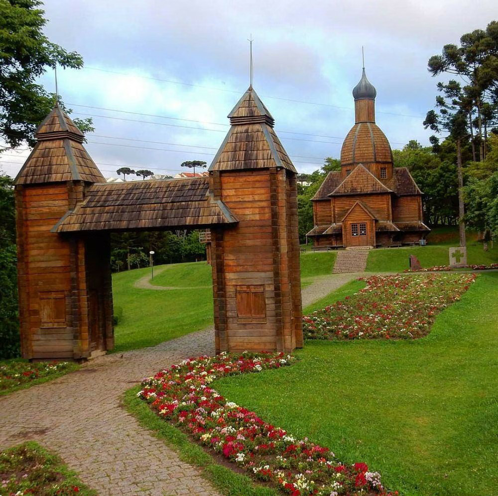 Wooden Ukrainian monument with domed towers and ornate gateway in landscaped park with flower beds