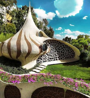Shell-shaped house with striped exterior, stone details, flowers, and green lawn under a blue sky