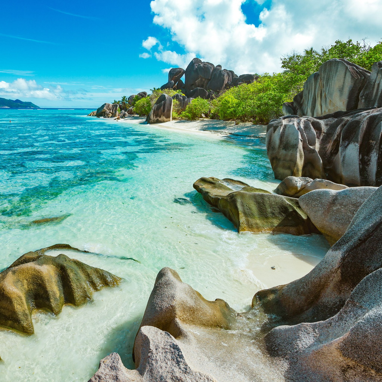 Tropical beach with large granite rocks, turquoise ocean, white sand, and green foliage under bright blue sky