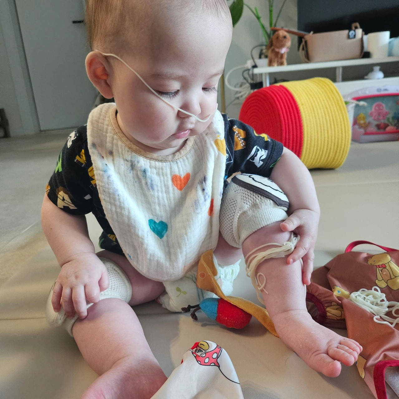 Baby sitting on the floor with spaghetti on their face, wearing a heart bib and surrounded by toys