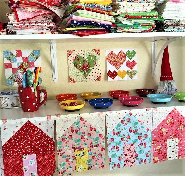 Sewing workspace with fabric stacks, quilt wall hangings, and colorful house-shaped fabric decorations