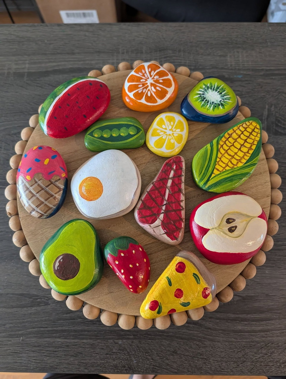 Painted rocks resembling fruits and foods arranged in a circle on a wooden board