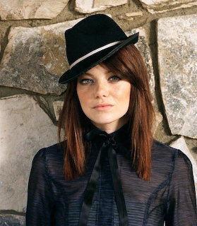 Woman with long reddish-brown hair wearing black fedora hat and dark blouse against stone wall