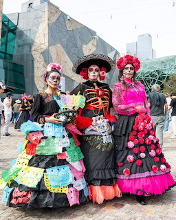 Three women in elaborate Day of the Dead costumes and skull makeup posing in a city square