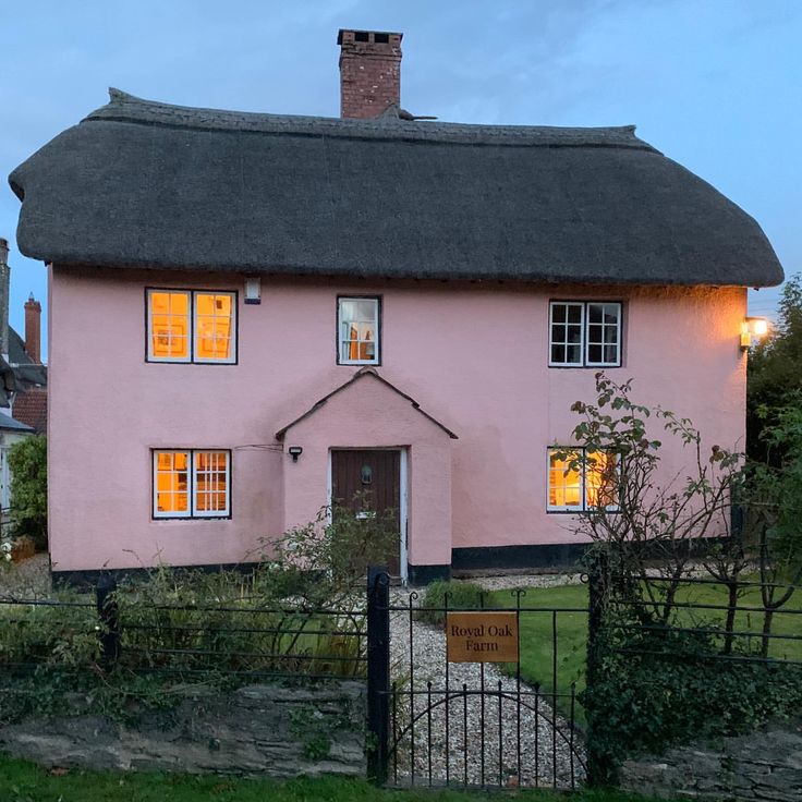 Pink thatched cottage with illuminated windows at dusk, garden path and gate in foreground
