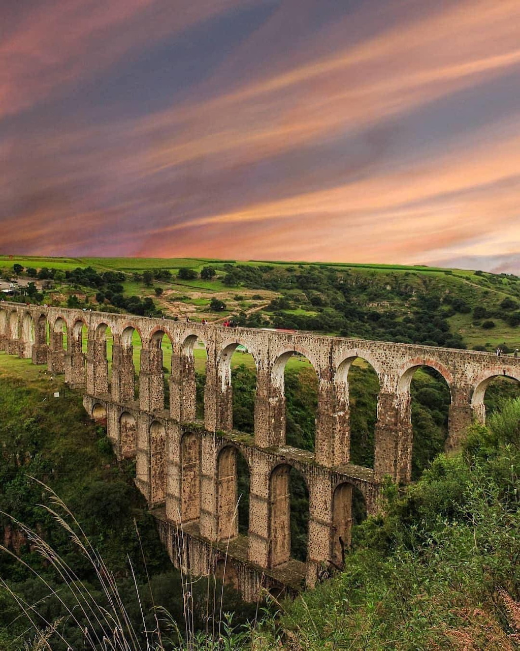 Old stone aqueduct with many arches crossing green hills under a colorful sunset sky