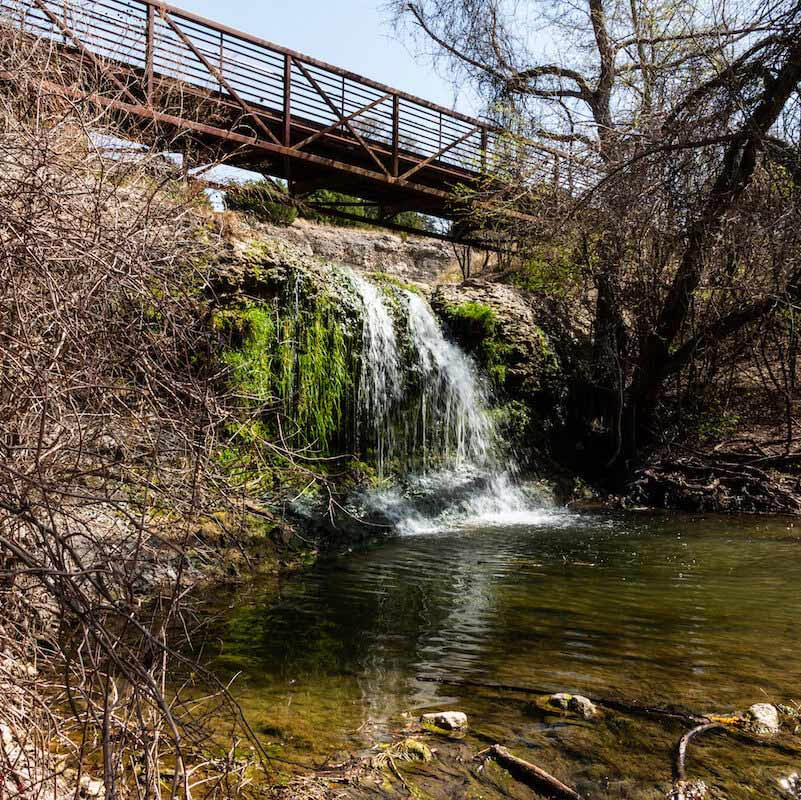 Small waterfall flowing into a pond beneath a metal bridge in a wooded area