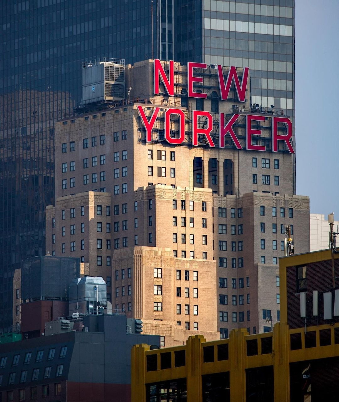 Beige New Yorker Hotel building with red neon sign, surrounded by glass skyscrapers at dusk