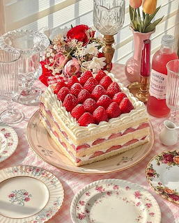 Square layered cake topped with fresh strawberries on decorative plate surrounded by vintage floral dishes and pink glassware