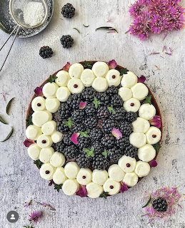Overhead view of berry tart with cream rosettes and dark berries on white wooden table with flowers