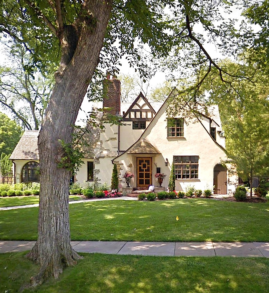 Tudor Revival style home with cream stucco walls, timber accents, green lawn, and large shade tree