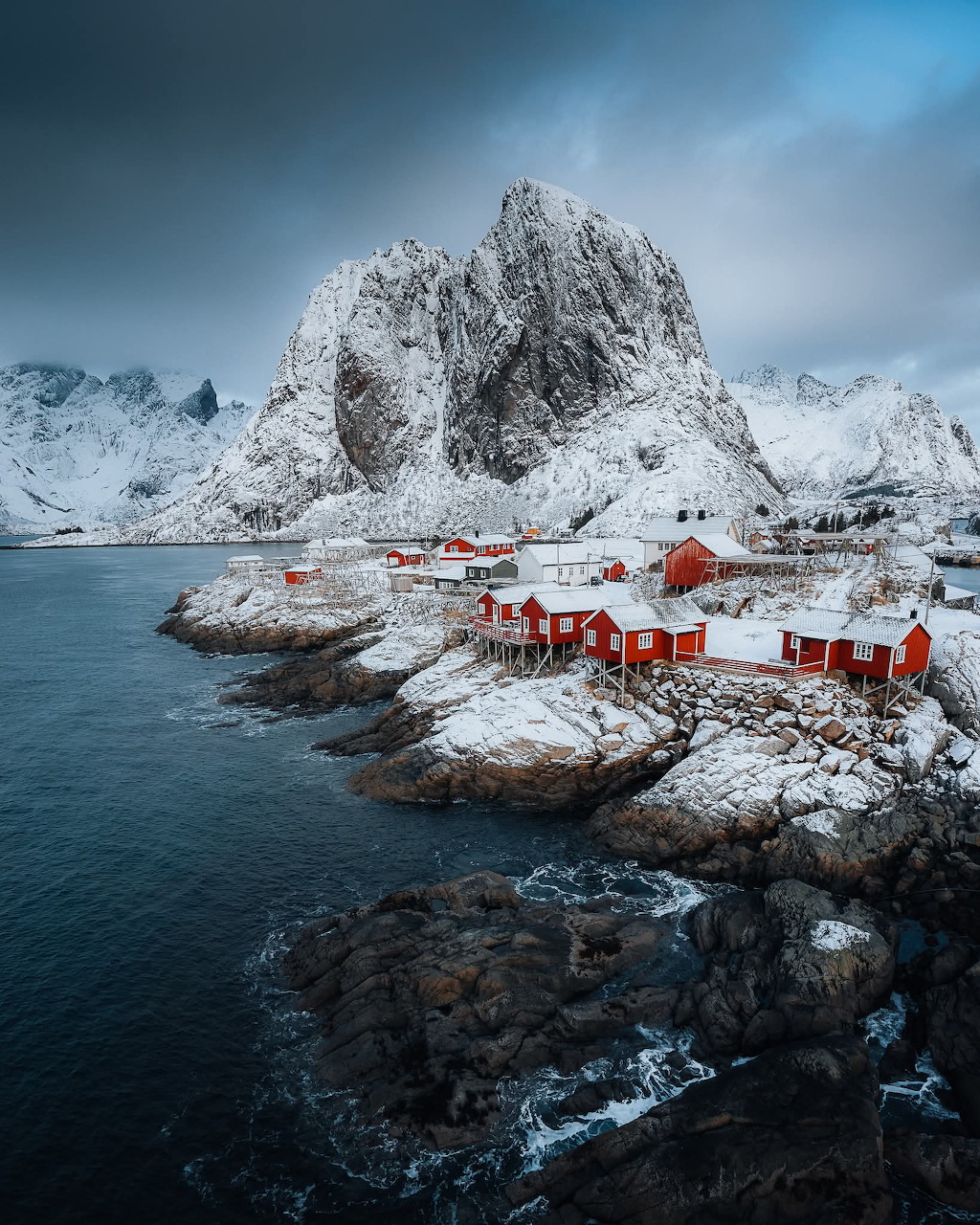Red fishing huts on snowy Lofoten coastline with steep snow-capped mountains and dark fjord waters
