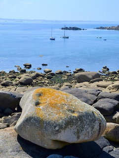 Rocky shore with large lichen-covered stones, calm blue sea and boats in background