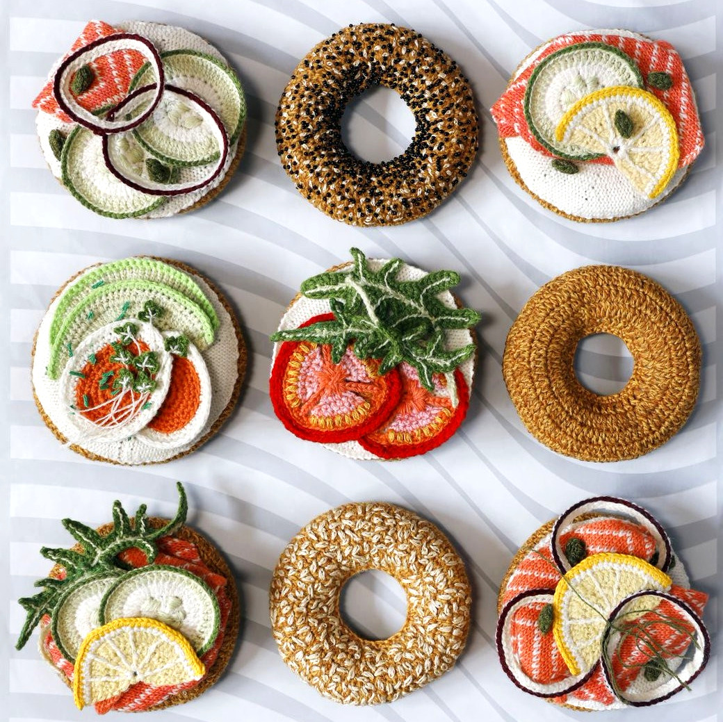 Nine crocheted bagels with toppings like salmon, tomato, cucumber, and cream cheese on white marble