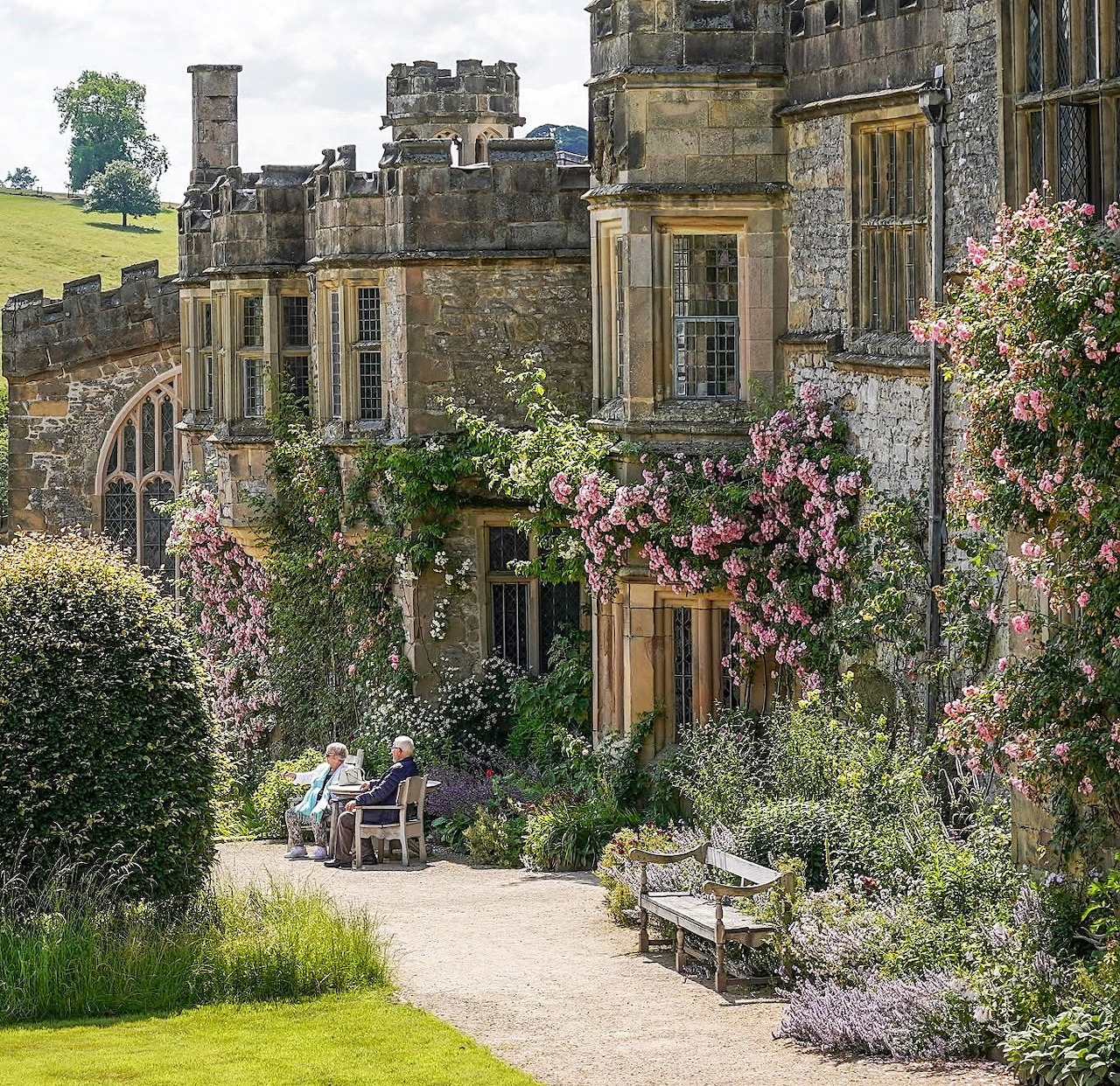 Stone medieval manor house with pink climbing roses and a garden path with visitors sitting outside
