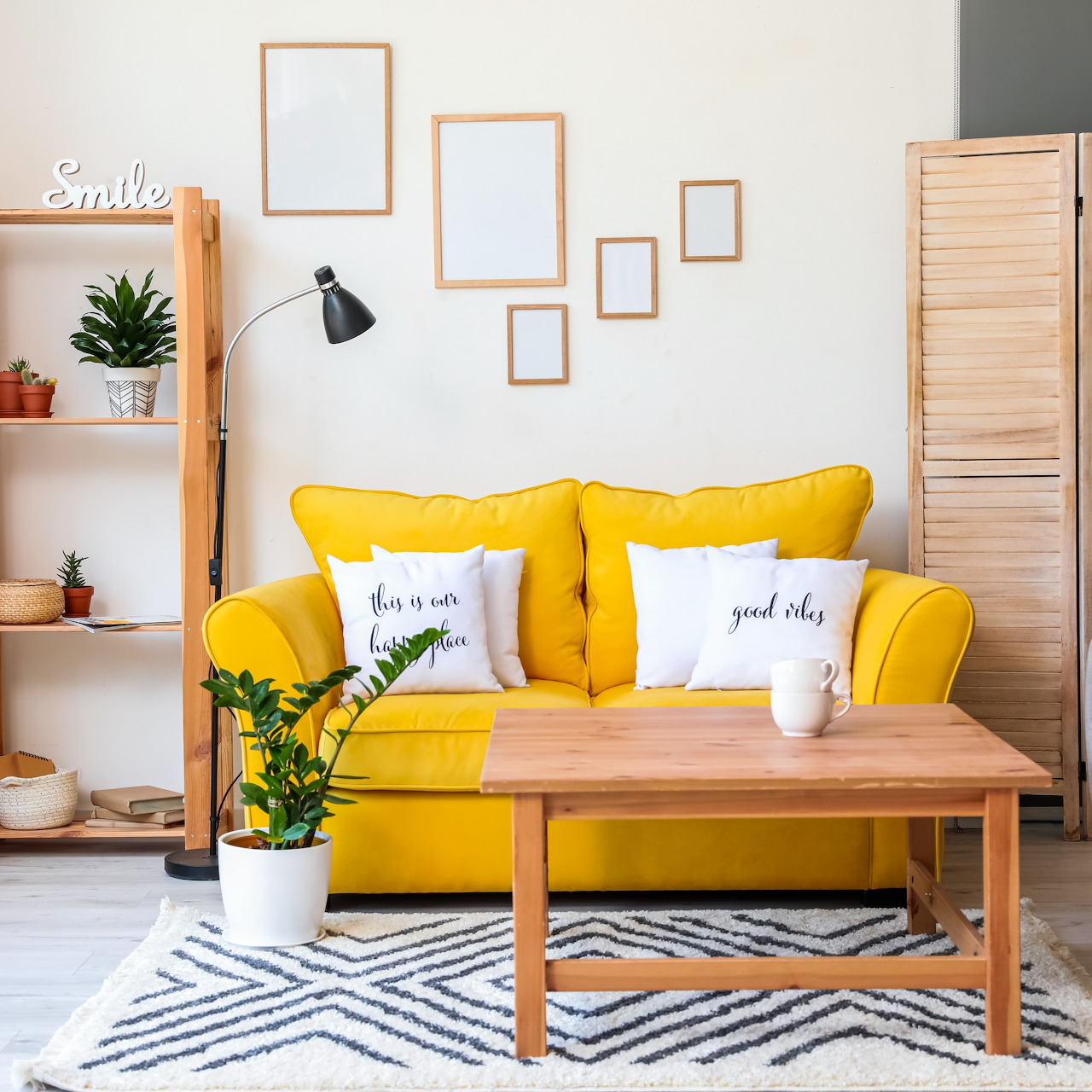 Modern living room with bright yellow couch, wooden table, geometric rug, empty picture frames on wall, and green plants