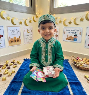 Young boy in green traditional clothing sitting on a blue mat with Eid Mubarak decorations and sweets