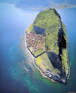 Aerial view of a green rocky peninsula with a small village on top surrounded by blue ocean