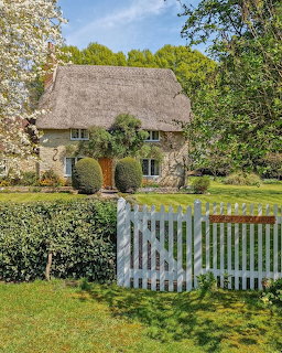 Thatched English cottage behind a white gate and hedge, surrounded by trees and green lawn