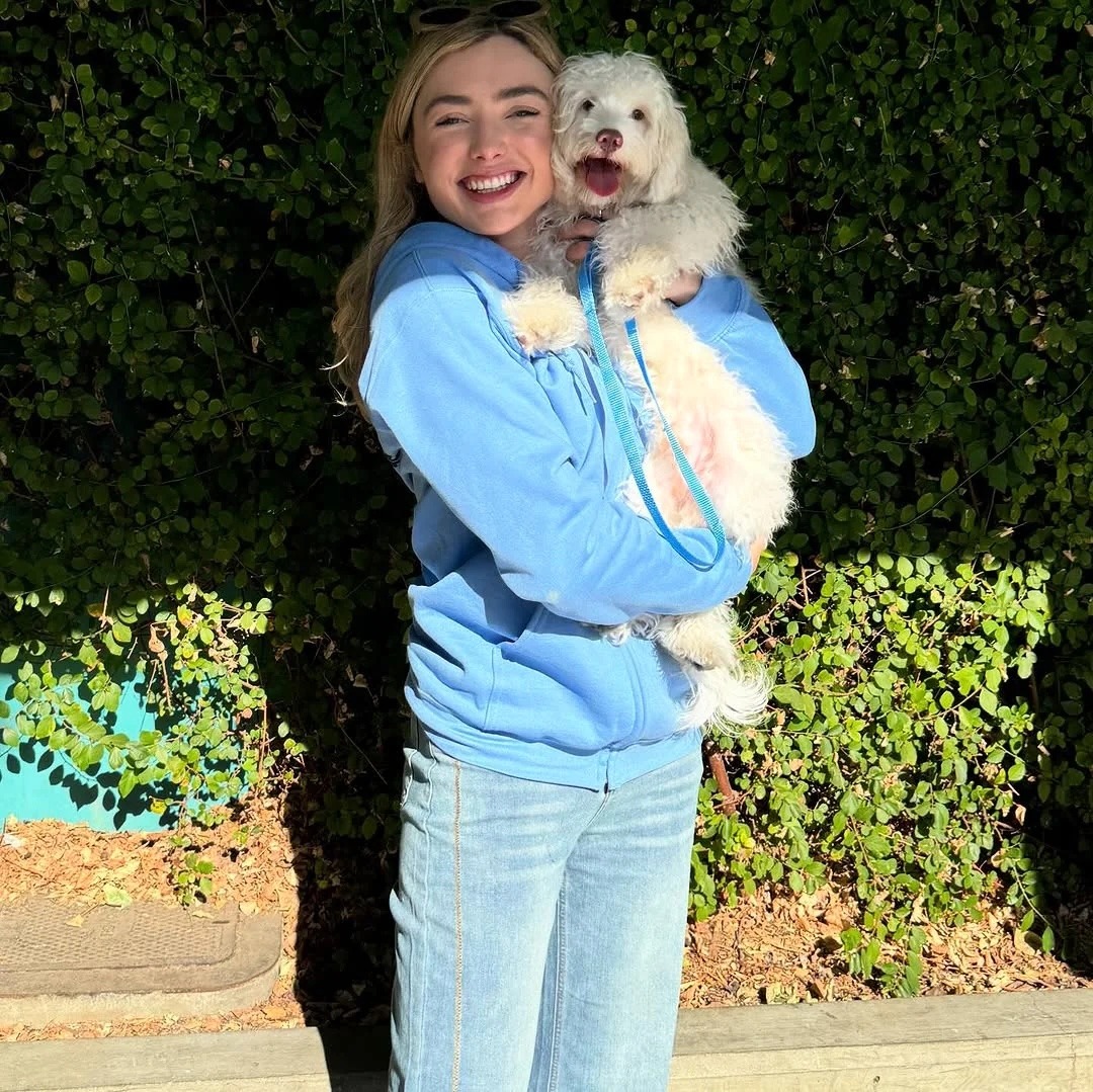 Smiling woman in light blue clothes holding a fluffy white dog outdoors by a green hedge