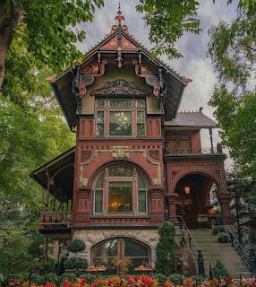 Victorian mansion with ornate carved details, bay windows, and flower gardens under tree canopy