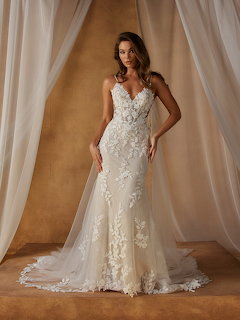 Bride wearing ornate white lace wedding dress with floral details and long train against draped fabric backdrop