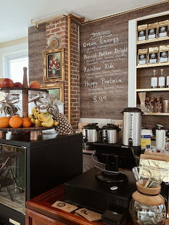 Coffee shop counter with chalkboard menu, fruit basket, coffee urns, and wooden shelves with jars