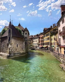 Medieval stone castle and tower next to canal with clear turquoise water, historic buildings lining waterway in Annecy