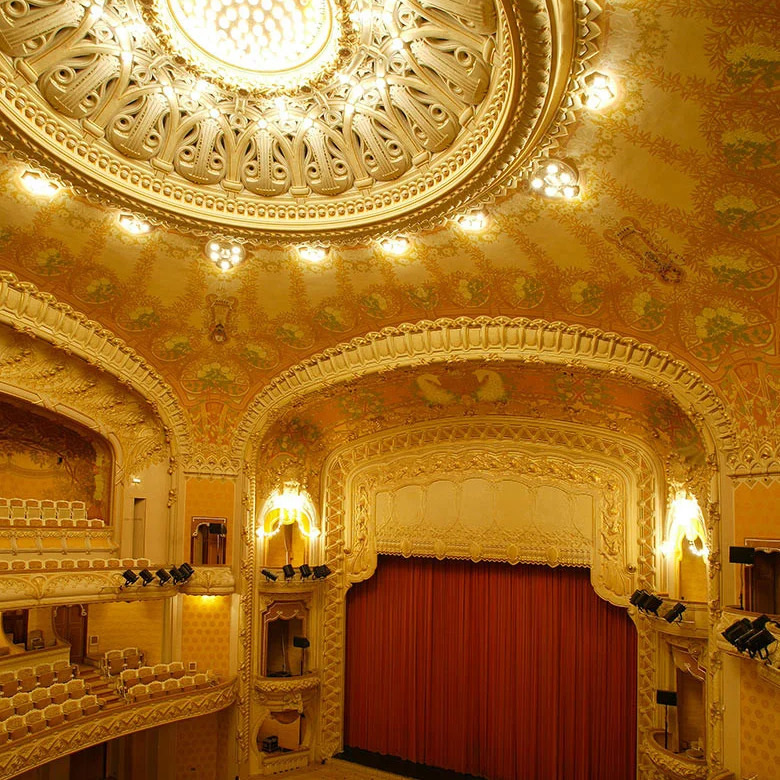 Ornate golden opera house interior with domed ceiling, decorative arches, and red curtain