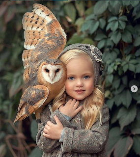 Young blonde girl holding a barn owl outdoors in front of leafy greenery