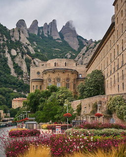 Montserat monastery with roundedapse and stone walls beneath jagged rocky peaks with flower gardens in front