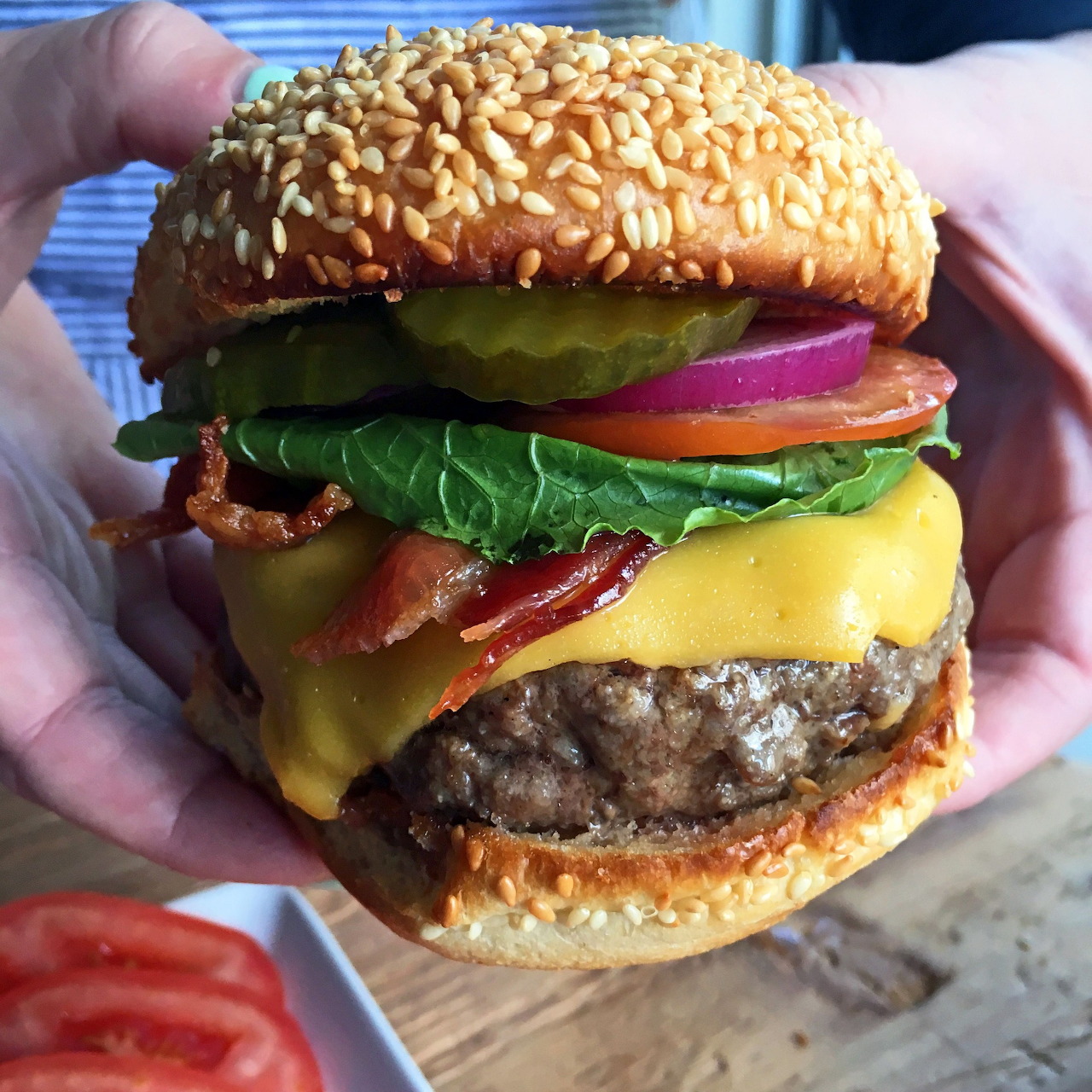 Hand holding a cheeseburger with bacon, lettuce, tomato, cheese, and sesame seed bun